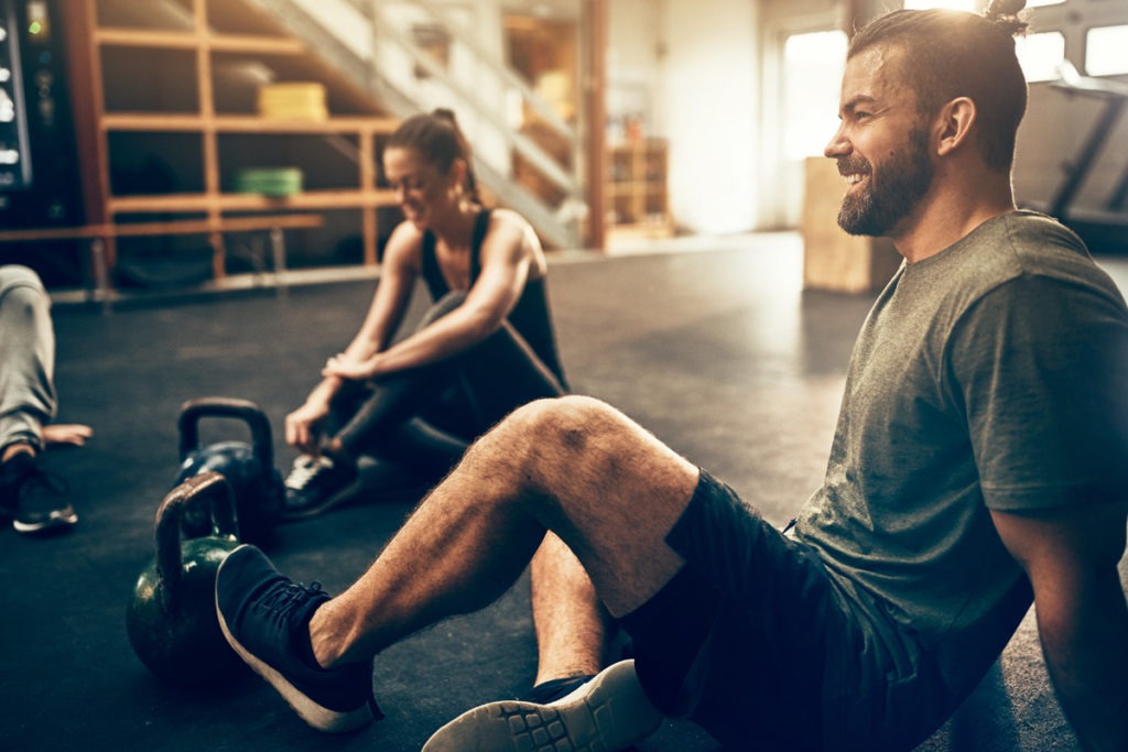 Fit people in exercise gear sitting on the floor of a gym laughing together after a workout